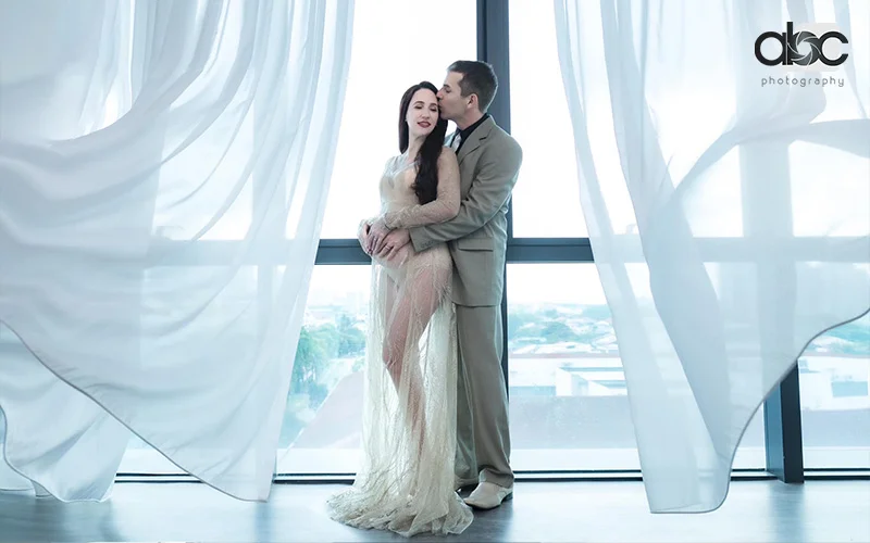 Couple embracing, sheer gown, light suit, flowing curtains, pregnancy photoshoot by abc photography